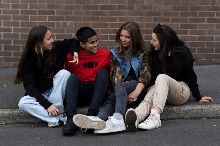 a group of teenagers of mixed genders sat on a curb chatting to each other.